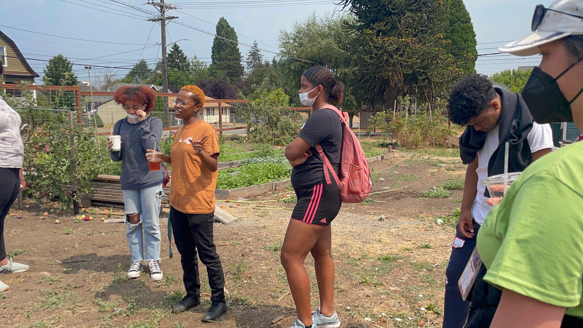 A young Black nonbinary person is speaking to a group of youth in a somewhat neglected urban farm